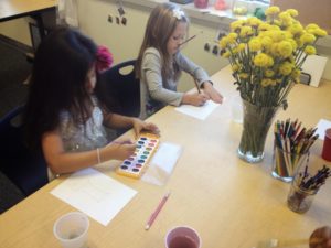 Two children painting with watercolors while looking at a vase of yellow flowers