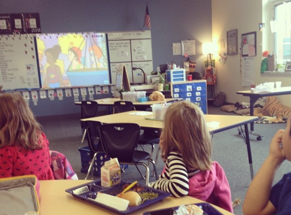 children eating lunch at a table while listening to a story in a classroom with a blue wall